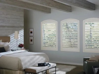A bedroom featuring three arched windows with white shutters, a bed with white bedding, a chair, and a beam ceiling.